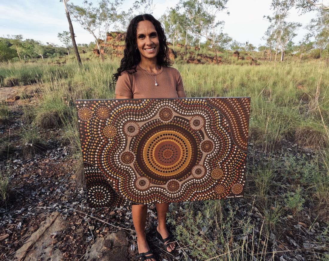 Taminga Connell standing on Country holding one of her paintings, concentric dotted circles in ochre, gold and brown.