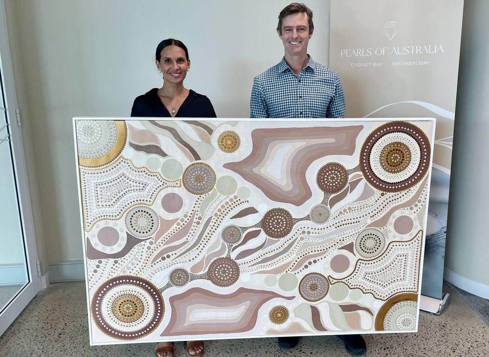 Taminga Connell and James Brown of Pearls of Australia holding a large commissioned painting in pearl tones, in front of a Cygnet Bay banner.